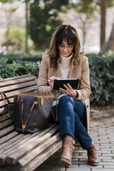 Ältere Frau, die ein digitales Tablet benutzt, während sie auf einer Bank im Park sitzt - EGAF02044