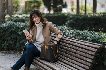 Mature woman using smart phone while sitting on bench at park - EGAF02046