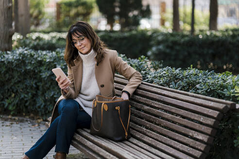 Reife Frau, die ihr Smartphone benutzt, während sie auf einer Bank im Park sitzt - EGAF02046