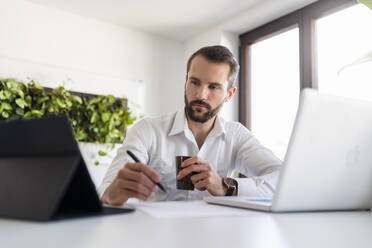 Geschäftsmann mit Laptop und Kaffeetasse bei der Nutzung eines digitalen Tablets im Büro - DIGF14823