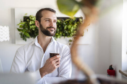 Thoughtful entrepreneur smiling while holding coffee cup at office - DIGF14825