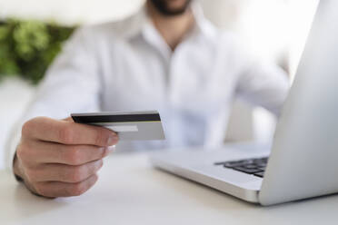 Entrepreneur with laptop holding credit card while sitting at desk in office - DIGF14832