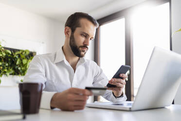 Berufstätiger mit Laptop und Kreditkarte bei der Nutzung eines Mobiltelefons im Büro - DIGF14833