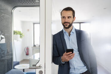 Junger Unternehmer mit Mobiltelefon an Telefonzelle im Büro gelehnt - DIGF14845