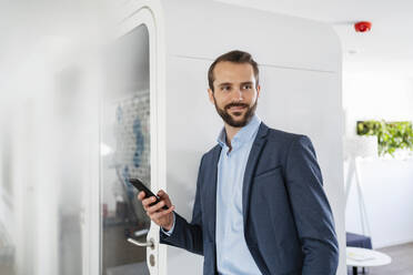 Geschäftsmann mit Mobiltelefon, der lächelnd an einer Telefonzelle im Büro steht - DIGF14847