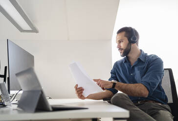 Male professional with headphones checking paper while using computer at office - DIGF14862