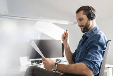 Male entrepreneur with headphones checking paper while sitting at office - DIGF14864