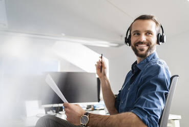 Smiling businessman with headphones and paper sitting at office - DIGF14865