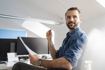 Junger Unternehmer mit Papier, der wegschaut, während er im Büro sitzt - DIGF14866