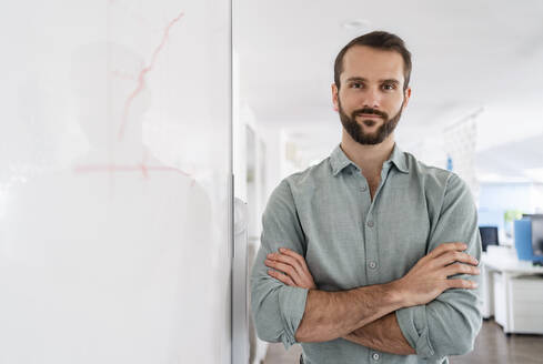 Confident businessman standing with arms crossed by whiteboard at office - DIGF14878