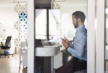 Businessman talking on video call over laptop while sitting in telephone booth at office - DIGF14897