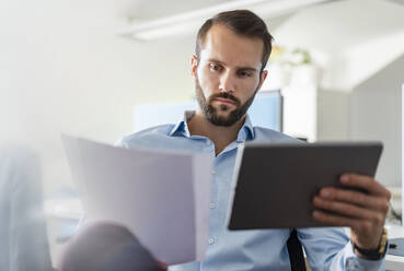 Geschäftsmann mit digitalem Tablet, der bei der Arbeit im Büro Papier analysiert - DIGF14902