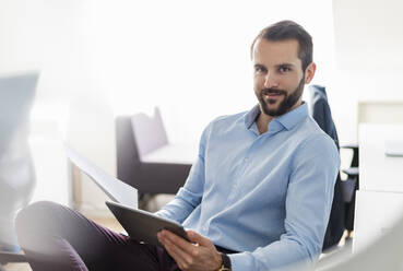 Confident businessman holding paper and digital tablet while sitting in office - DIGF14906