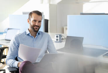 Lächelnder Unternehmer mit digitalem Tablet und Papier im Büro sitzend - DIGF14907