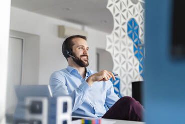 Nachdenklicher Unternehmer mit Kopfhörern, der lächelnd im Büro sitzt - DIGF14909