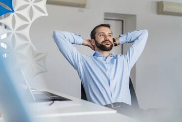 Young businessman with hands behind head sitting at office - DIGF14910