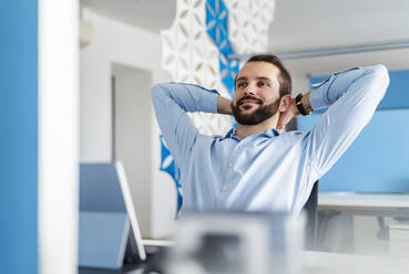 Thoughtful businessman smiling while sitting with hands behind head at office - DIGF14911