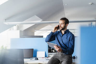Berufstätiger mit digitalem Tablet, der im Büro sitzend mit seinem Mobiltelefon spricht - DIGF14919