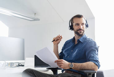 Smiling businessman with headphones and paper sitting at office - DIGF14928