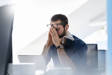 Young entrepreneur with protective face mask sitting with head in hands at office - DIGF14930