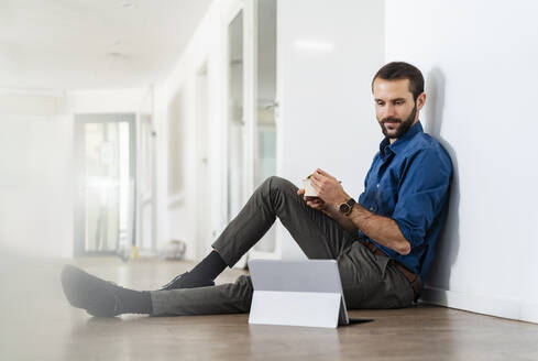 Junger Geschäftsmann schaut auf ein digitales Tablet, während er im Büro etwas isst - DIGF14935
