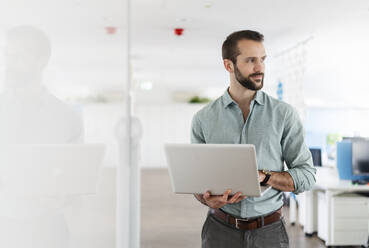 Young professional with laptop looking away while standing at office - DIGF14941