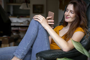 Relaxed young woman using smart phone while sitting on chair at home - AFVF08453