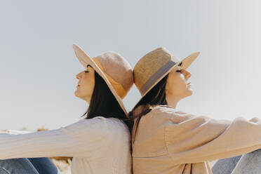 Young sisters back to back sitting by clear sky on sunny day - ARTF00046