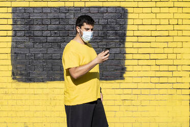 Mann mit Schutzmaske, der sein Smartphone benutzt, während er während einer Pandemie an einer gelben Wand steht - FBAF01622