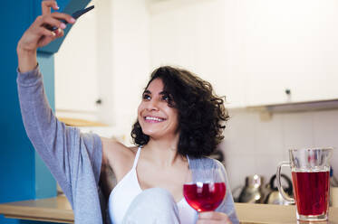 Smiling woman holding glass of juice while taking selfie through mobile phone at kitchen - PGF00466