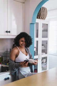 Lächelnde Frau, die eine Obstschale hält, während sie ihr Smartphone in der Küche benutzt - PGF00467