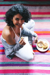 Smiling woman having breakfast while sitting on floor at home - PGF00468