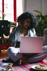 Frau mit Hand im Haar, die einen Laptop benutzt, während sie zu Hause auf dem Boden sitzt - PGF00471