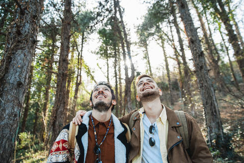Cheerful gay couple looking up while hiking in forest during vacations - DAF00018