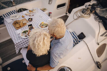 High angle view of senior couple taking selfie on mobile phone in sailboat during sunset - MASF22298