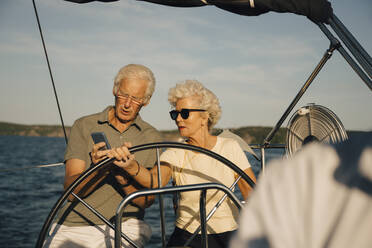 Älteres Paar, das beim Segeln in einem Boot an einem sonnigen Tag mit dem Smartphone die Richtung angibt - MASF22330