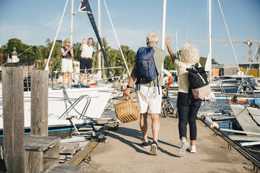 Senior friends waving at heterosexual couple while walking on pier at harbor - MASF22359