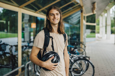 Porträt eines lächelnden männlichen Studenten mit Fahrradhelm an einer Fahrradabstellanlage auf dem Campus - MASF22459