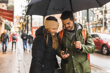 Lächelndes Paar, das ein Smartphone benutzt, während es auf einem Fußweg in der Stadt während der Regenzeit spazieren geht - MASF22460