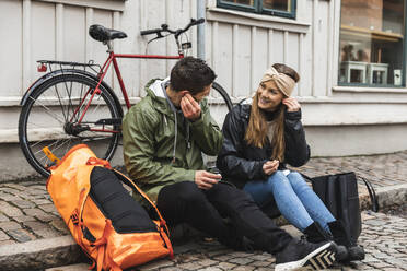 Männliche und weibliche Touristen tragen In-Ear-Kopfhörer, während sie auf einem Fußweg in der Stadt sitzen - MASF22464