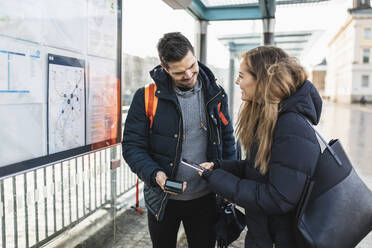 Lächelnde Frau, die einen QR-Code mit ihrem Smartphone scannt, neben einem Mann am Bahnhof stehend - MASF22493