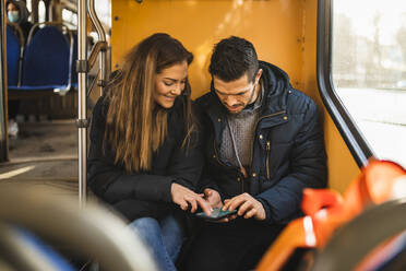 Heterosexuelles Paar, das während einer Pandemie in der Straßenbahn sitzt und ein Smartphone benutzt - MASF22494