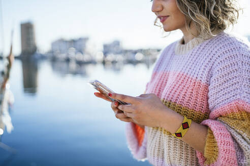 Lächelnde Frau in warmer Kleidung, die ein Mobiltelefon am Fluss benutzt - MPPF01556