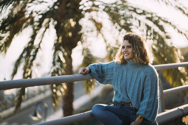 Lächelnde Frau in warmer Kleidung, die wegschaut, während sie im Frühling am Geländer sitzt - MPPF01560