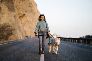 Smiling woman with Siberian Husky and Jack Russell Terrier looking away while walking on mountain road against sky - MPPF01566