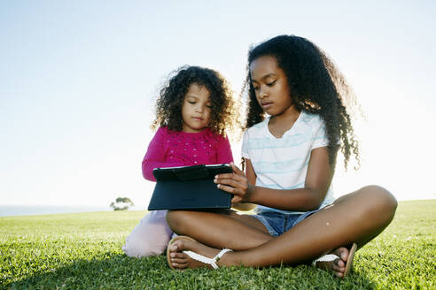 Junges gemischtrassiges Mädchen und ihre jüngere Schwester sitzen im Freien und teilen sich ein digitales Tablet - MINF15938
