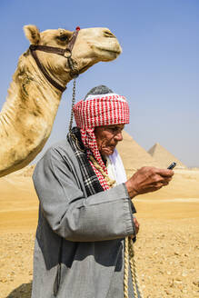 Ein Führer in traditioneller arabischer Kleidung, Gewand und Turban, der ein Mobiltelefon benutzt, und sein Kamel. - MINF16007