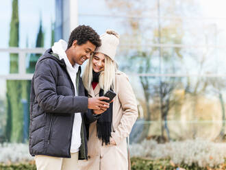 Junge aufrichtige multiethnische Partner in warmer Kleidung, die auf ihr Handy schauen, während sie gemeinsam Zeit in der Stadt verbringen - ADSF21085