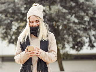 Junge fröhliche Frau in warmer Kleidung surft im Internet auf dem Handy in der Stadt an einem sonnigen Tag - ADSF21087