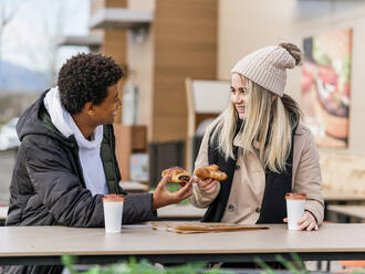 Junge, fröhliche, multiethnische Freunde mit köstlichem Gebäck und Einweggläsern mit Getränken, die sich in einem Straßencafé unterhalten und einander ansehen - ADSF21088